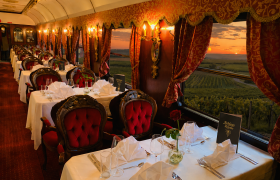 Eleganter Speisewagen eines Zuges mit rot-goldener Einrichtung, gedeckten Tischen und gepolsterten St&uuml;hlen; durch die gro&szlig;en Fenster ist eine Landschaft mit Weinbergen bei Sonnenuntergang zu sehen.