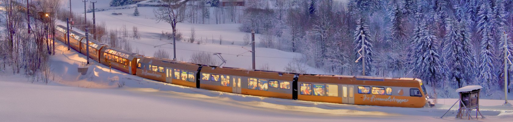 Gelb-orangefarbener Zug fährt durch verschneite Winterlandschaft mit schneebedeckten Bäumen und Gleisen.