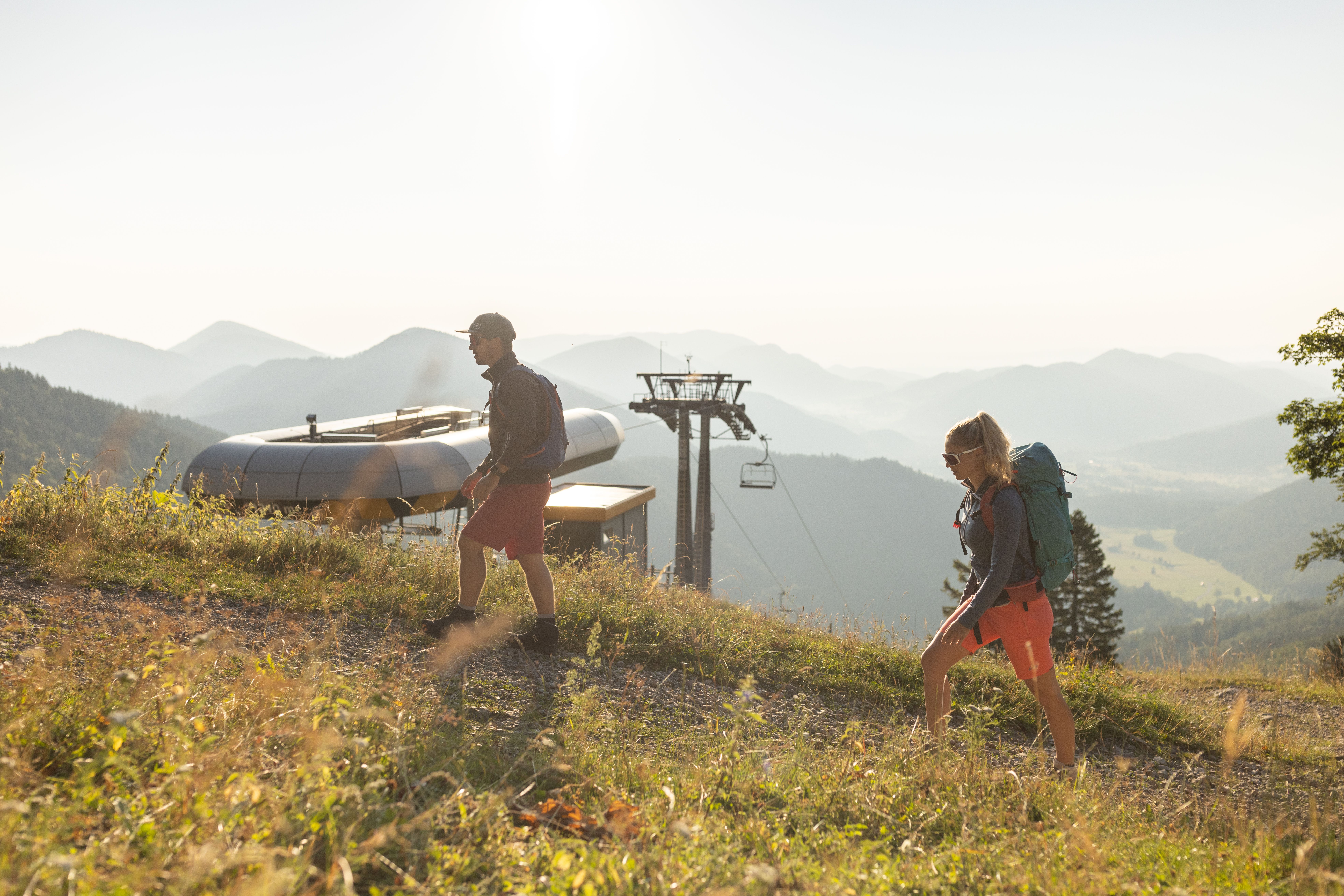 Zwei Wandernde mit Rucksäcken gehen an der Bergstation der Schneeberg Sesselbahn vor einem weitläufigen Bergpanorama bergauf.