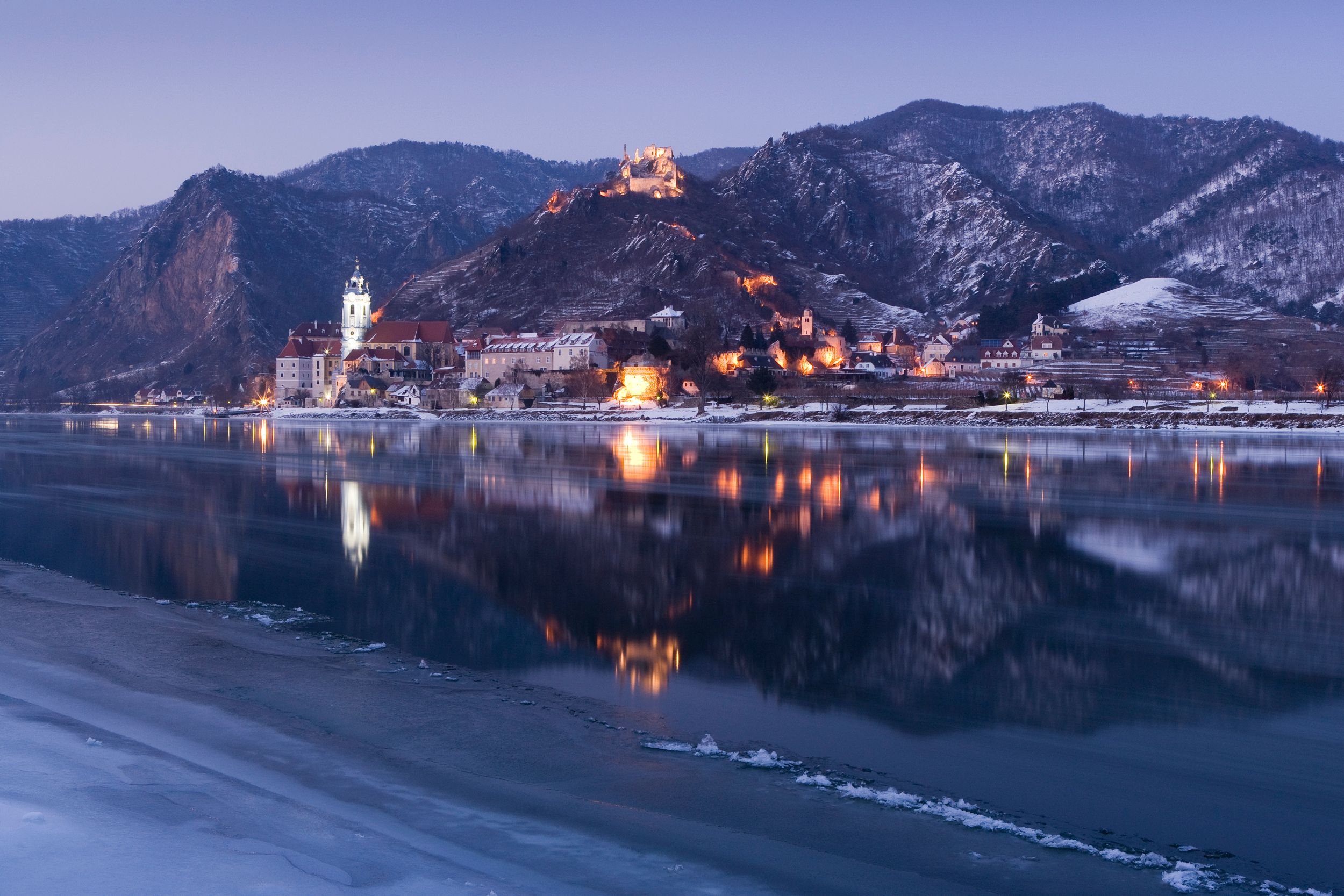 Winterliche Ansicht von Dürnstein an der Donau mit beleuchteten Gebäuden und schneebedeckten Hügeln im Hintergrund.