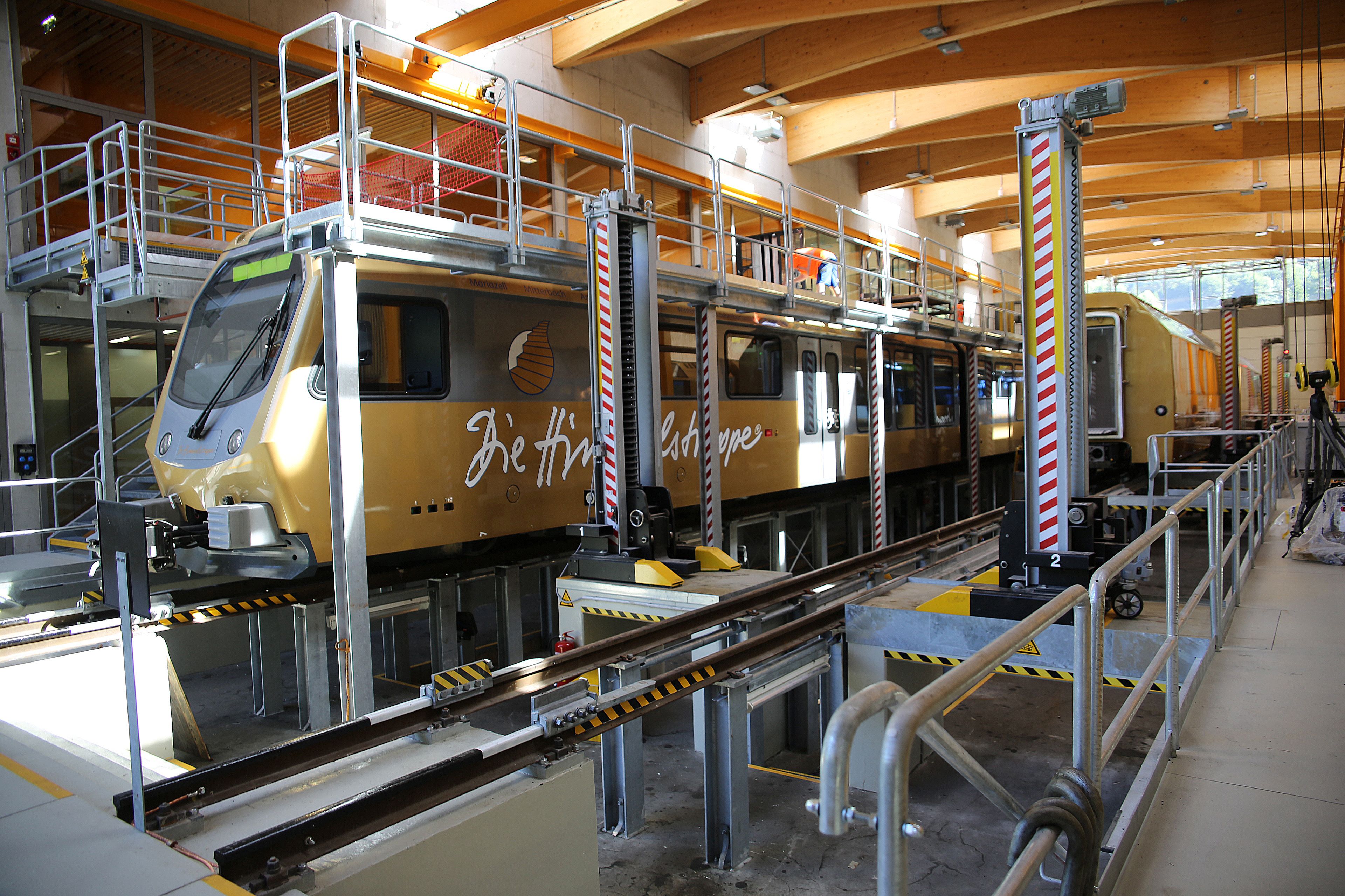 Goldfarbener Triebwagen der Mariazellerbahn steht in einer modernen Werkstatthalle auf einem Wartungsgleis, umgeben von Hebeanlagen und Arbeitsbühnen.