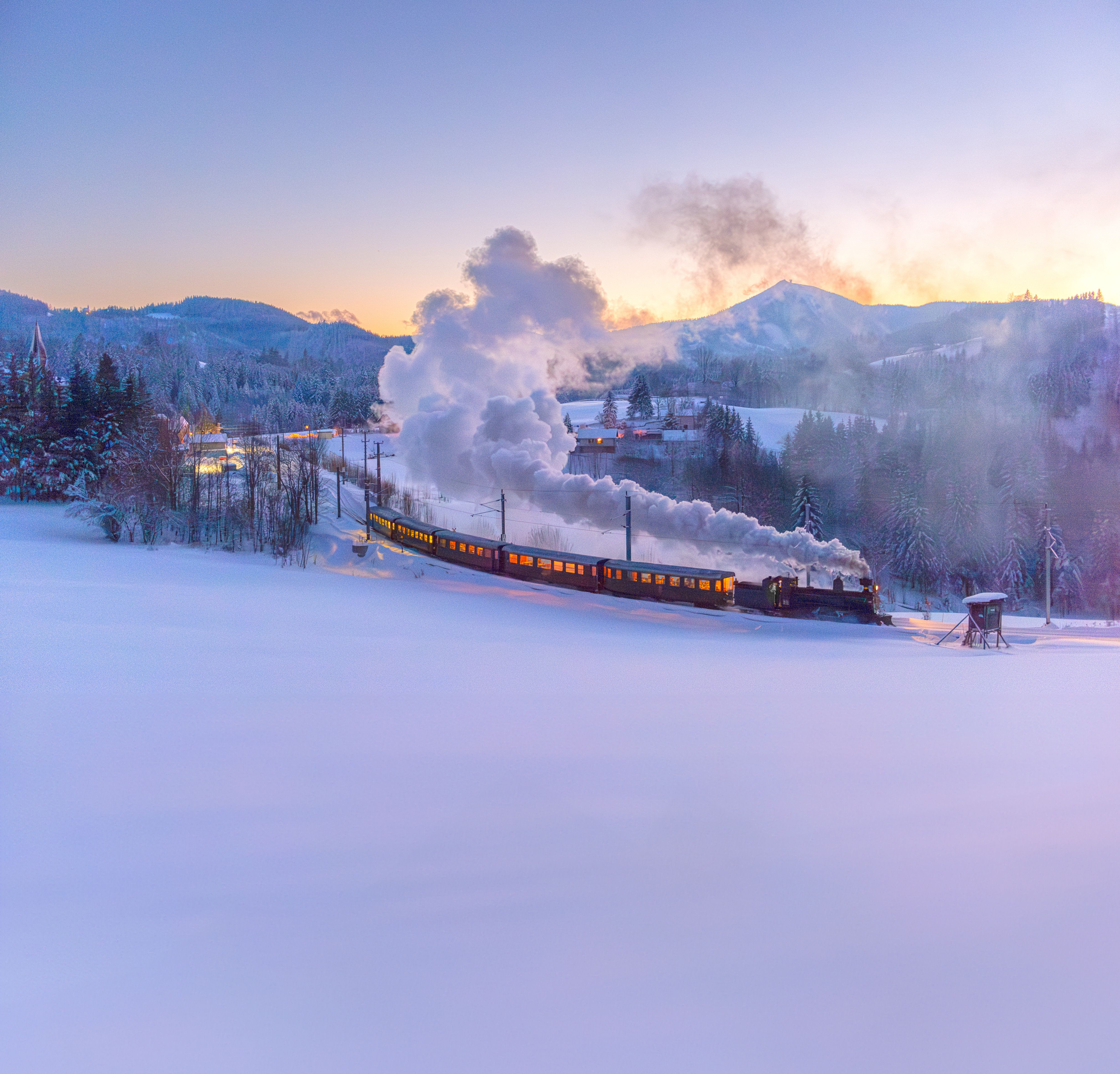Dampfzug fährt durch eine tief verschneite Winterlandschaft. Beleuchtete Waggons und dichte Dampfwolken heben sich vor den bewaldeten Bergen im Abendlicht ab.