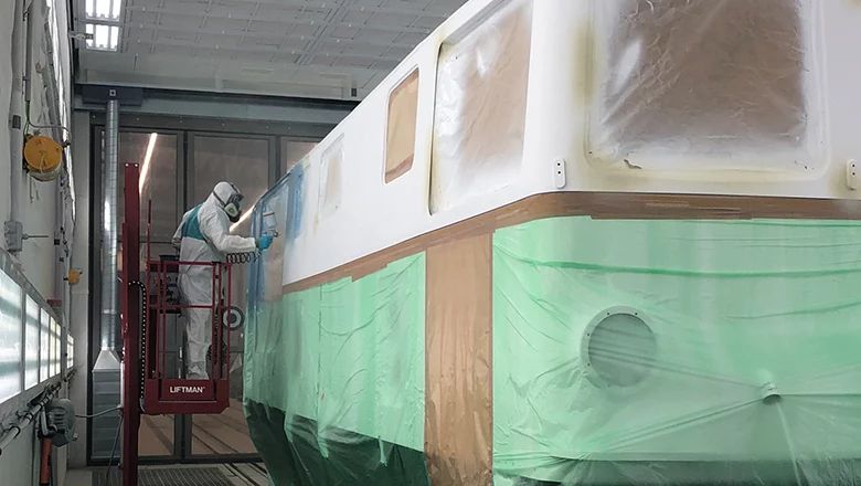 Mitarbeiter in Schutzanzug und Atemmaske lackiert in einer Werkstatt die Seite eines Schienenfahrzeugs. Fenster und Fahrzeugteile sind sorgfältig abgedeckt.