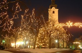 Weihnachtlich beleuchteter Marktplatz mit Bäumen, die mit Lichterketten geschmückt sind, und einer beleuchteten Kirche im Hintergrund bei Nacht