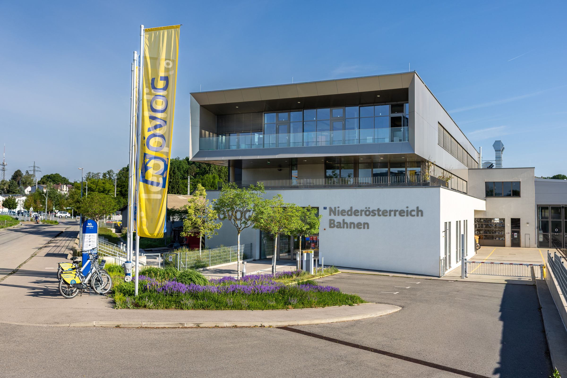 Modernes mehrstöckiges Gebäude mit Glasfront und gelber Fahne mit Aufschrift "NÖVOG" vor blauem Himmel