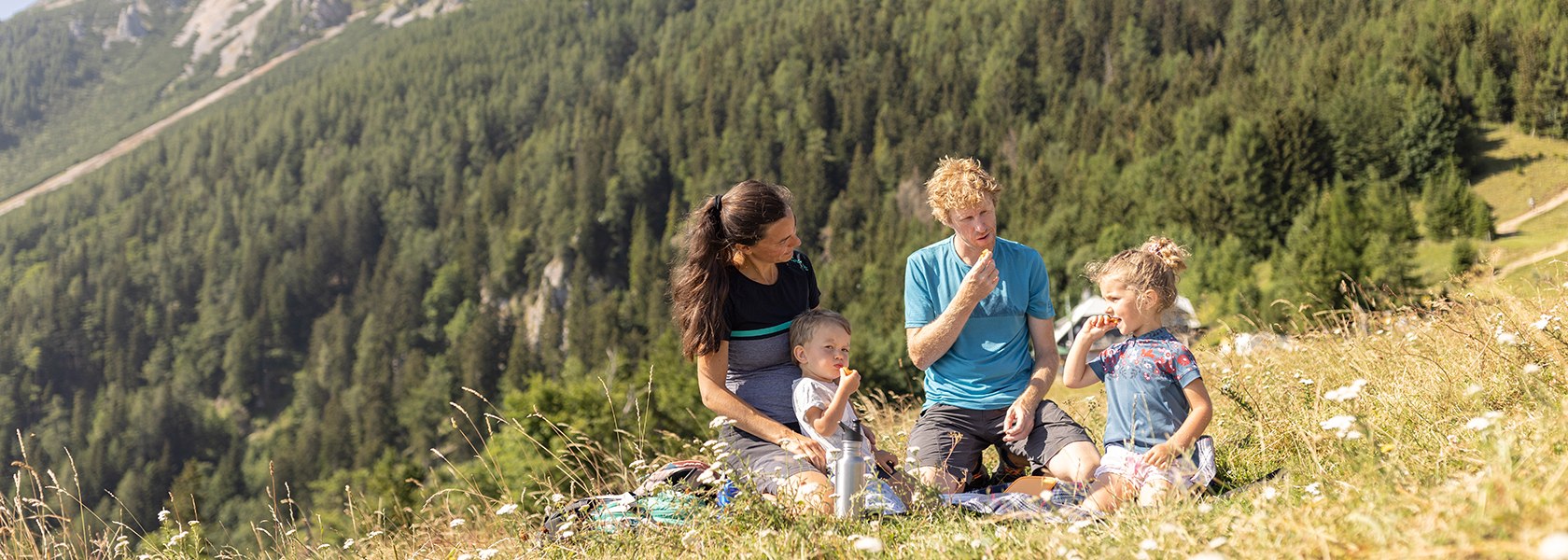 Familie sitzt auf blühender Wiese mit Picknickdecke und Snacks