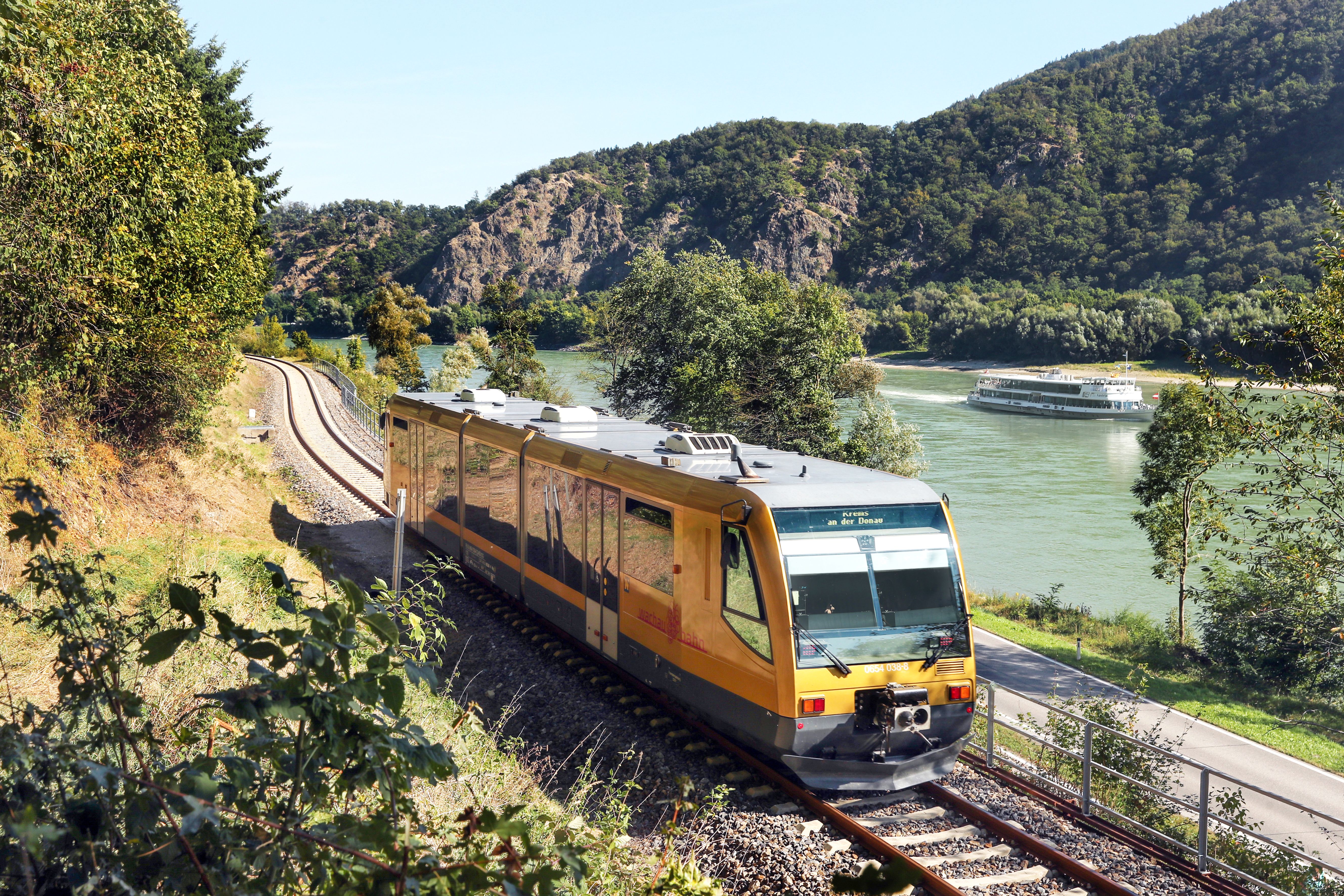 Goldene Wachaubahn fährt entlang der Donau; auf dem Fluss ist ein Ausflugsschiff unterwegs.