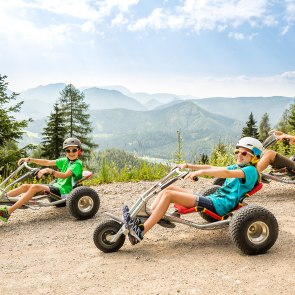 Bursch, Mädchen und die Großmutter genießen eine Mountaincart-Abfahrt. Im Hintergrund sieht man das Bergpanorama