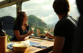 Zwei Personen sitzen an einem Tisch im Zug und sto&szlig;en mit Weingl&auml;sern an, im Hintergrund sind Berge durch das Fenster zu sehen.