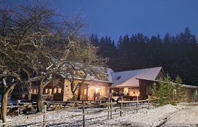 Beleuchtetes Gasthaus mit schneebedecktem Dach und Boden, umgeben von Bäumen und Tischen im Freien bei Abenddämmerung