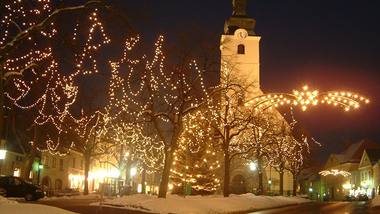 Weihnachtlich beleuchteter Marktplatz mit Bäumen, die mit Lichterketten geschmückt sind, und einer beleuchteten Kirche im Hintergrund bei Nacht
