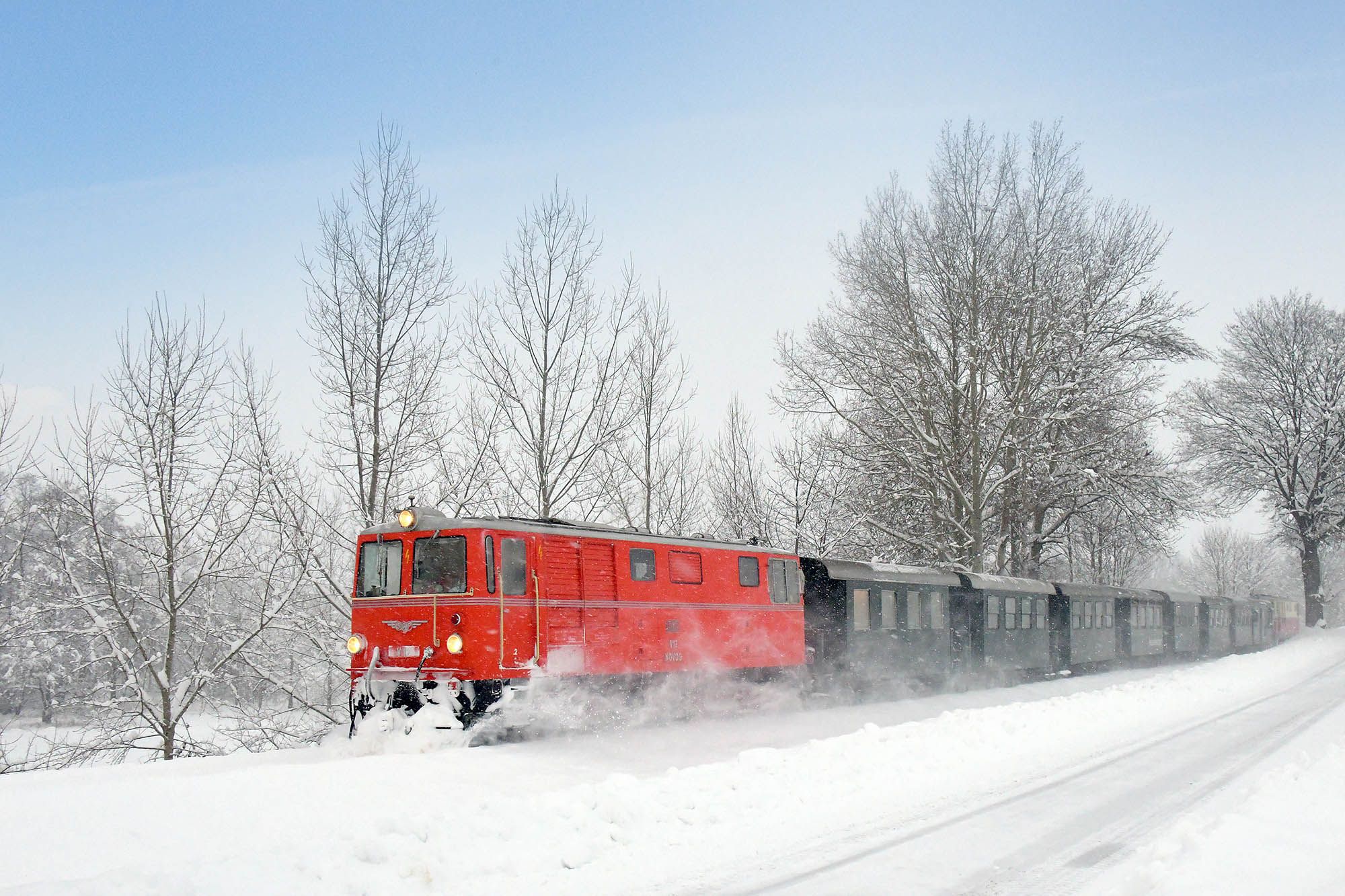 Roter Zug fährt durch verschneite Landschaft mit kahlen Bäumen.